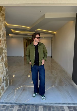 Chaqueta verde oliva de botones con bolsillos en el pecho, camiseta negra, jeans amplios azules y zapatillas verde y blanco sobre un suelo de mármol.