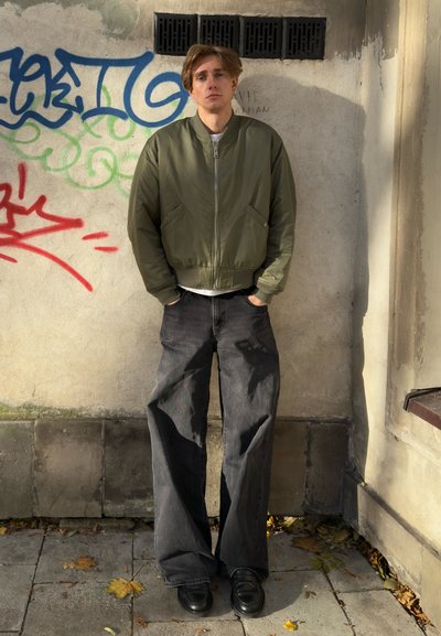 Chaqueta bomber verde oliva con cremallera frontal, jeans negros de pierna ancha y zapatos negros. Fondo con grafiti en una pared texturizada.