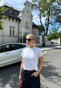 Bela krajšane majica, temno visoko pasje kavbojke in sončna očala; stoji ob belem avtu in svetli stavbi z okrasnimi detajli.