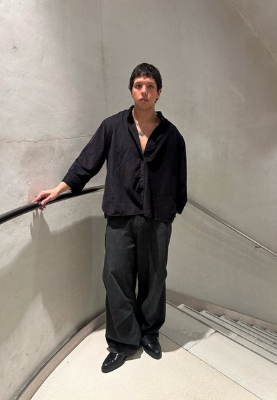Camisa de lino negra con cuello abierto y corte holgado, combinada con pantalones anchos a rayas negras y zapatos negros, contra un fondo minimalista.