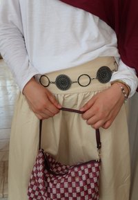 Ręce trzymają czerwoną torebkę w kratkę z łańcuszkiem, ubrane w białą koszulę z długim rękawem, beżową spódnicę, ozdobny pasek i bransoletki.