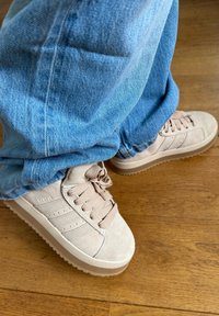 Ljust beige mockasins i mocka med strukturerade, rundade kanter och tre ränder på sidan; ihop med blå denimbyxor på ett trägolv.
