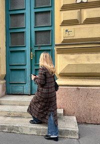 Karirani dugi kaput u smešnim i crnim tonovima, uparen s plavim farmerkama. Žena se penje sivim kamenim stepenicama prema tirkiznim dvostrukim vratima s staklenim panelima.