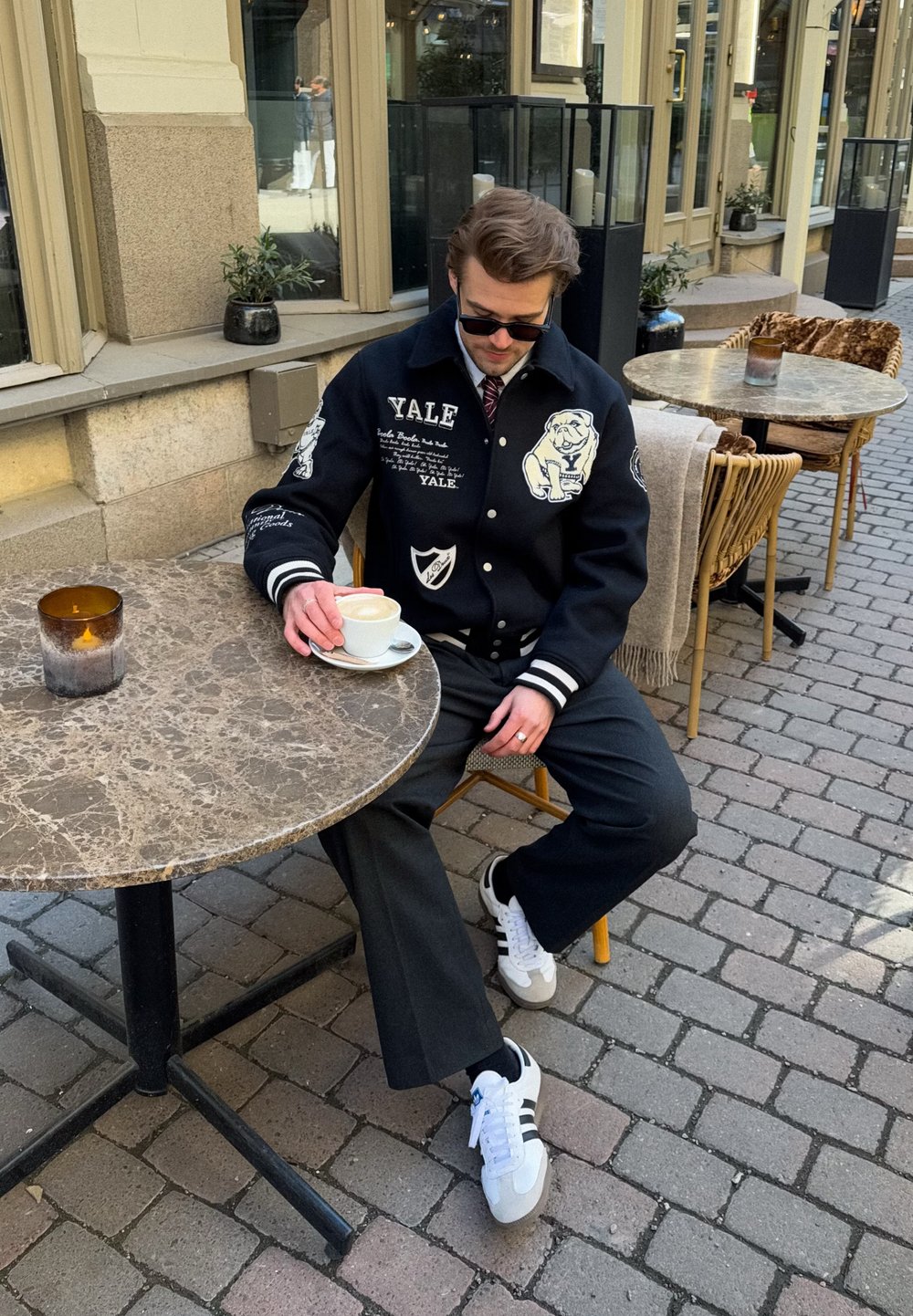 Casaco varsity preto com texto e gráficos de Yale, calças cinzentas e ténis brancos com riscas pretas. Sentado à mesa de mármore ao ar livre.