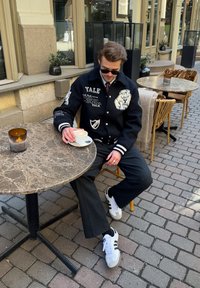 Casaco varsity preto com texto e gráficos de Yale, calças cinzentas e ténis brancos com riscas pretas. Sentado à mesa de mármore ao ar livre.