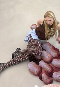 Un top maro, o fustă albastră și colanți cu plasă cu un model de diamant. Pantofi cu toc înalt negri. Împrejur sunt forme colorate de covorașe moi.