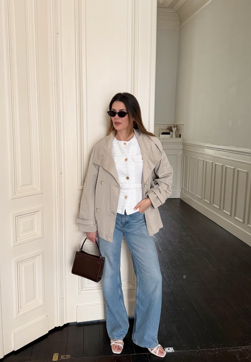 Woman wearing beige jacket, white buttoned shirt, wide-leg jeans, white sandals, black sunglasses, and holding a dark brown handbag standing indoors.