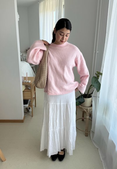 Suéter de lana rosa claro y suave con mangas largas, combinado con una falda larga blanca de volantes. El modelo sostiene un bolso tejido beige y lleva zapatos negros.