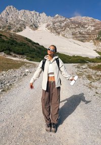 Jachetă ușoară crem peste un top alb scurt, asortată cu pantaloni cargo maro largi și bocanci gri de drumeție. Ține o sticlă de apă albă.