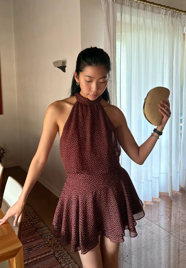 Donna in abito marrone senza maniche a pois, che tiene una clutch beige, in piedi all'interno vicino a un tavolo di legno e tende bianche trasparenti.