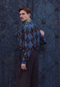 Homme avec une moustache portant un pull à losanges bleu et marron et un pantalon sombre, se tenant devant une porte métallique bleu foncé texturée.