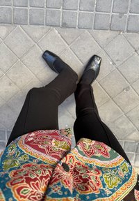 Personne portant un pantalon noir, des chaussures noires à bouts carrés et une veste colorée à motifs, se tenant sur un trottoir en dalles gris.