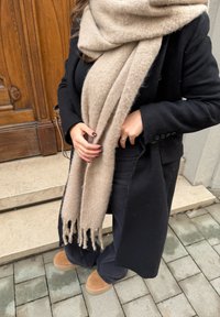 Μπεζ μάλλινο κασκόλ με φούντες, φορεμένο πάνω από ένα μαύρο παλτό. Μαύρα παντελόνια και ταμπά σουέτ μπότες είναι ορατά. Το φόντο περιλαμβάνει λίθινες σκάλες και ξύλινη πόρτα.