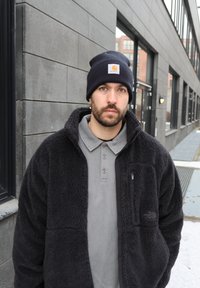 Habeme ja musta mütsiga mees, kes kannab fliisjakki, seisab talvel väljas halli hoone ja suurte akende ees.