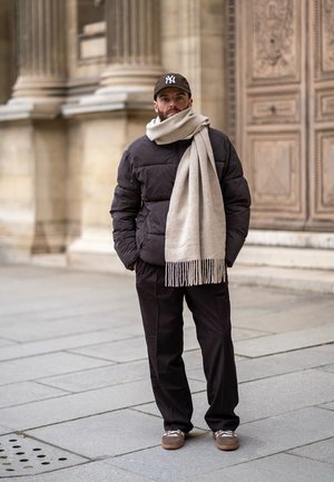 Chaqueta acolchada de color marrón oscuro, bufanda de tejido beige con flecos, pantalones negros y zapatillas marrones con cordones blancos. Gorra de béisbol oscura.