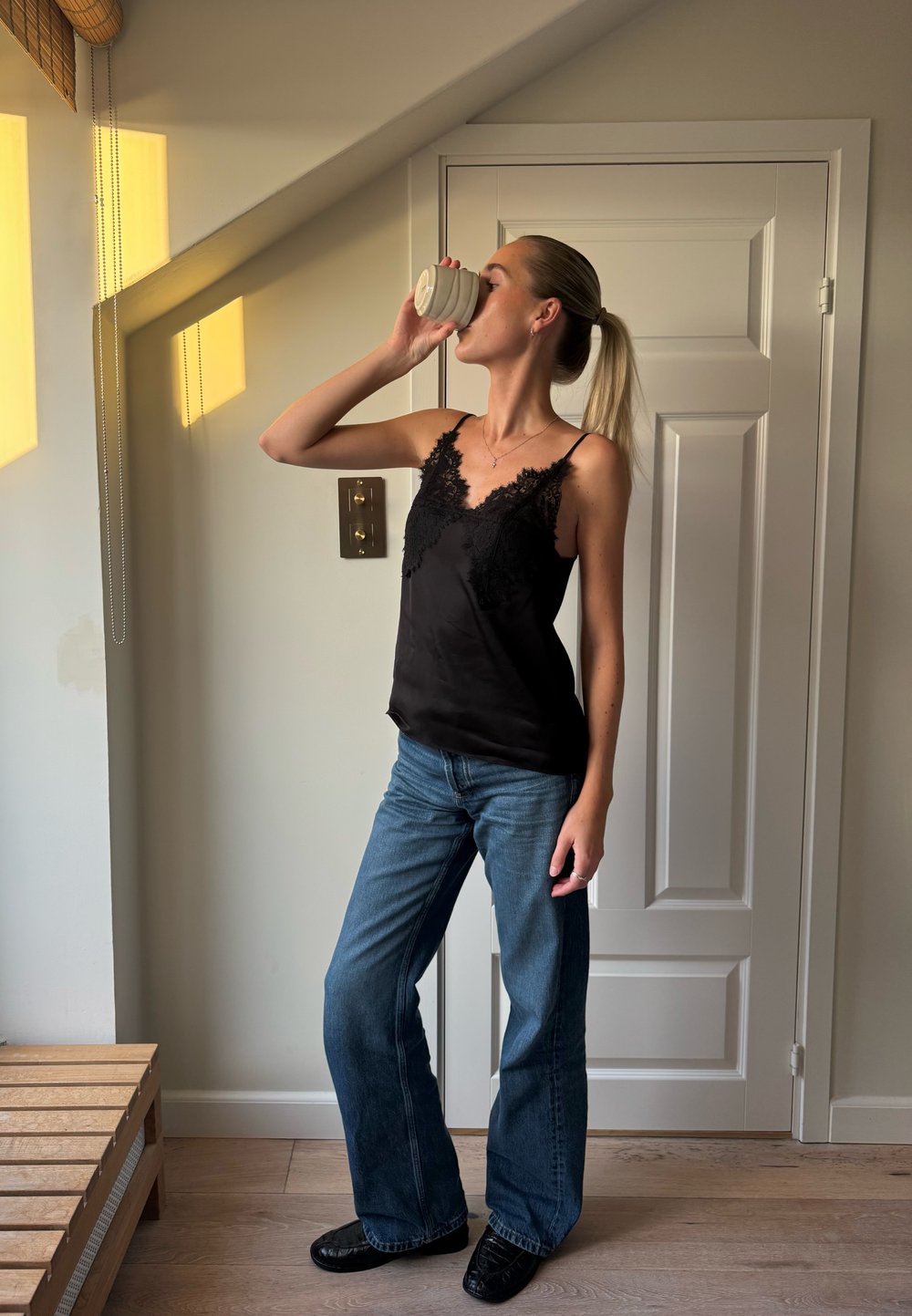 Camisola de cetim preta com borda de renda, combinada com jeans azuis claros flare. A pessoa segura uma caneca branca, com uma parede neutra ao fundo.