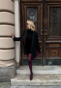 Black blazer over a black polo neck, paired with burgundy tights and heels. Standing on steps against wooden paneled doors with ornate designs.