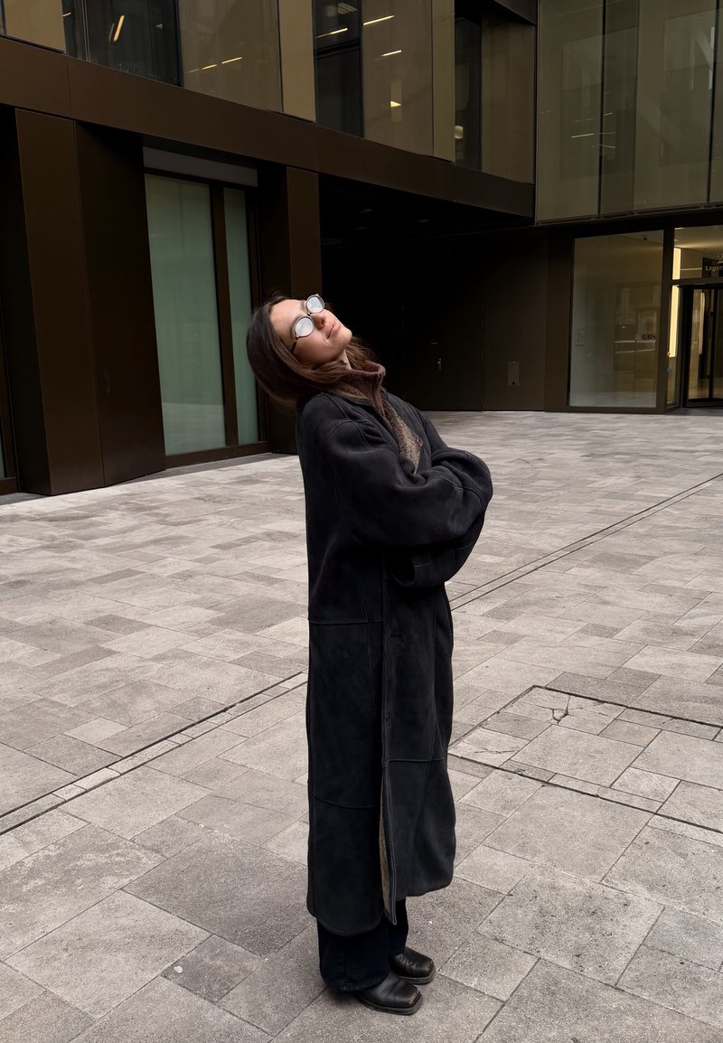 Mujer con abrigo largo negro y gafas, de pie con los brazos cruzados y la cabeza inclinada hacia atrás, en el patio de un edificio moderno con suelo de baldosas.