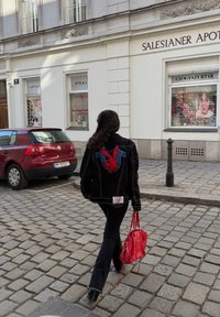 Casaco de ganga preto com o logótipo vermelho da Playboy, calças pretas e uma bolsa vermelha. Rua de calçada e lojas ao fundo.