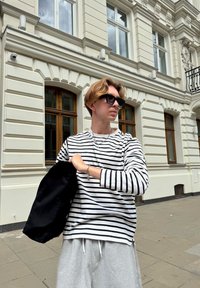 Prúžkovaná dlhá rukávová košeľa v čiernej a bielej farbe, šedé teplákové nohavice, drží čiernu tašku. Pozadie zobrazuje bielu budovu s drevenými oknami.