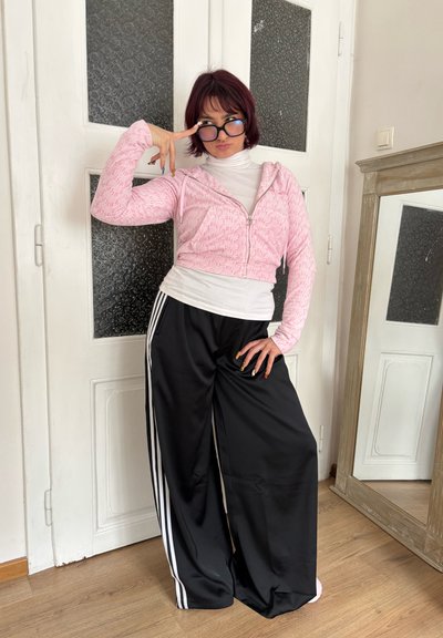 Sudadera corta rosa con cremallera y patrón texturizado, camiseta blanca de manga larga debajo y pantalones deportivos negros holgados con rayas blancas.