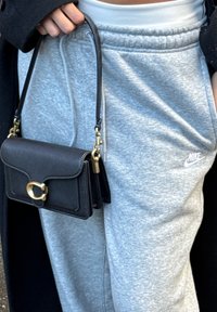 Černá kožená kabelka se zlatou sponou a popruhem přes rameno. Nošena s šedými bavlněnými tepláky. Viditelný texturovaný povrch.