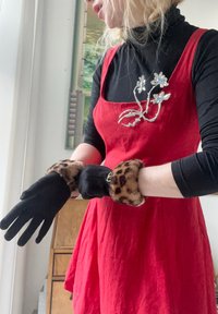 Mănuși negre cu manșete din blană cu imprimeu leopard, purtate cu o rochie roșie și un pulover negru cu guler înalt. O broșă florală din argint accentuează ținuta.