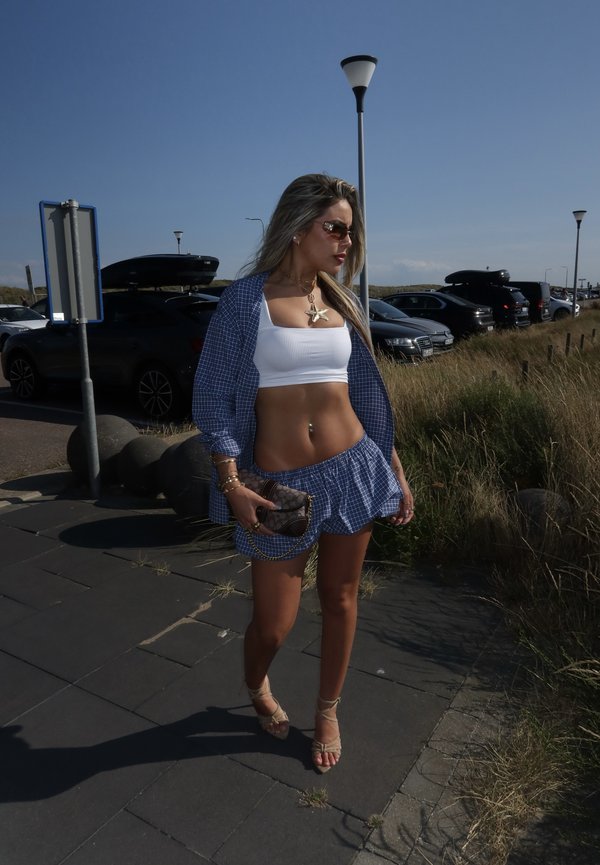 Femme portant un crop top blanc, un short bleu à carreaux et une veste, tenant un sac à main, debout sur un trottoir près de voitures garées et de l'herbe.