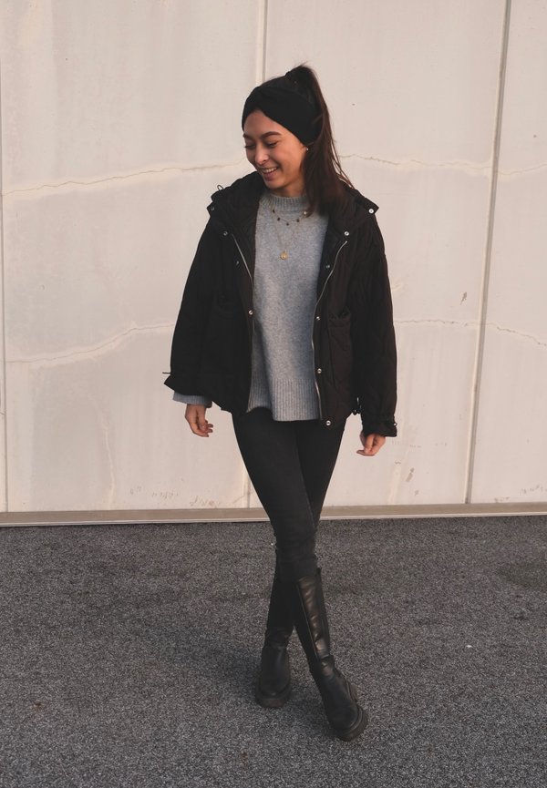 Black quilted jacket, gray knit sweater, black skinny jeans, and knee-high black boots. Background is light-colored with textured flooring.