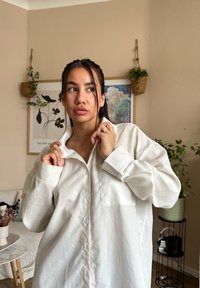 Λευκό πουκάμισο με κουμπιά, που διαθέτει μπροστινή τσέπη, μακριά μανίκια και άνετη εφαρμογή. Λείο ύφασμα με ελαφριά γυαλάδα, φορεμένο με τον γιακά ελαφρώς ανασηκωμένο.