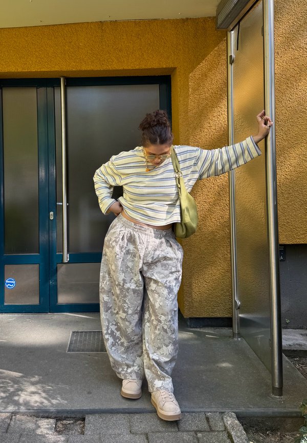 Femme en chemise rayée à manches longues et pantalon ample à motifs appuyée contre un poteau argenté devant un bâtiment jaune texturé avec une porte bleu foncé.