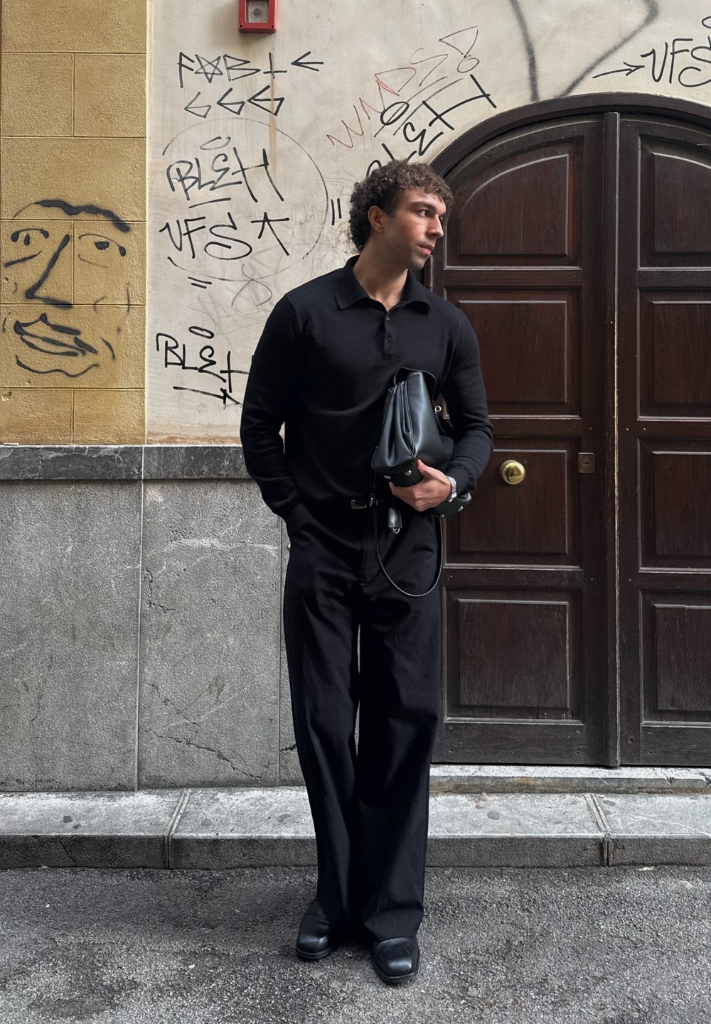 Homme portant une chemise noire à manches longues et un pantalon large tenant un sac noir, debout près d'un mur avec des graffitis et une porte en bois sombre.