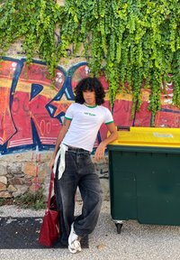 Λευκή μπλούζα με πράσινες λεπτομέρειες, φαρδιά μαύρα τζιν, κόκκινη τσάντα, λευκά αθλητικά παπούτσια; στέκεται δίπλα σε έναν πράσινο κάδο και μπροστά σε έναν ζωντανό τοίχο γκράφιτι.