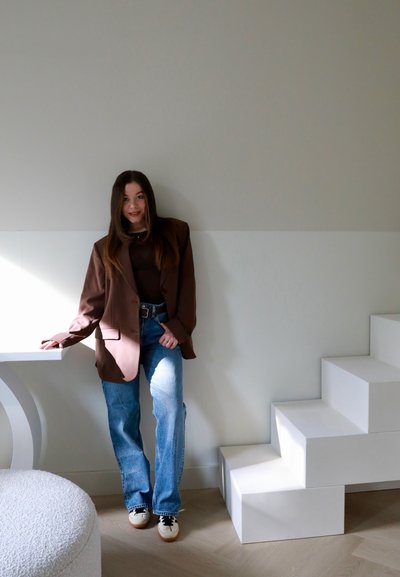 Blazer marrón de gran tamaño, top marrón ajustado, jeans rectos de pierna azul claro y zapatillas blancas. De pie junto a unas escaleras modulares blancas y una mesa.