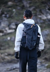 Persoon die een wit jack en zwarte broek draagt, met een zwarte rugzak en handschoenen, staand op een rotsachtig buitenpad.