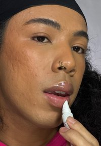 Baume à lèvres dans un applicateur blanc, appliqué sur les lèvres. Gros plan montrant la peau texturée, des lèvres pleines, un maquillage naturel et une chemise rose vif.