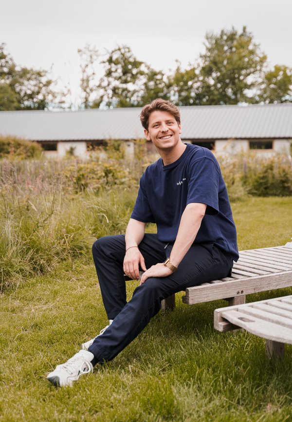 Camiseta de algodón color marino y pantalones negros, sentado en un banco de madera en un área con hierba. Zapatillas deportivas de color claro. Fondo natural con vegetación.