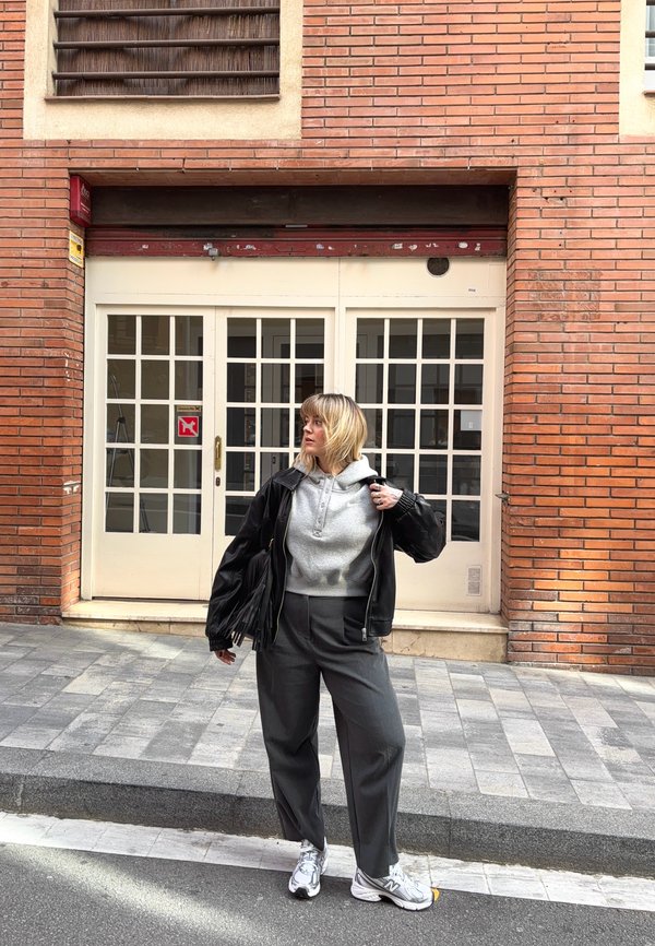 Woman with blonde hair stands on pavement wearing grey hoodie, black leather jacket, dark trousers, and white trainers in front of brick building with glass doors.