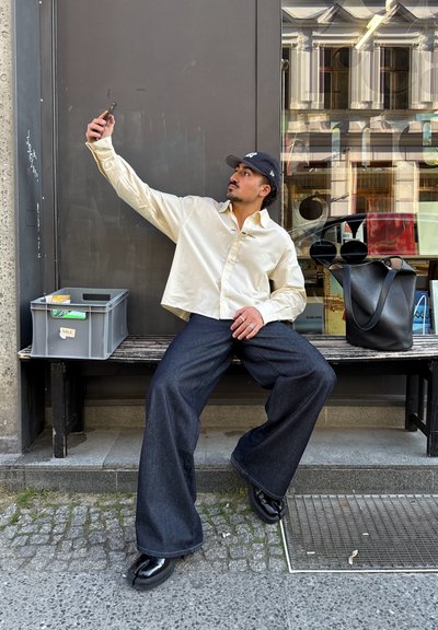 Una persona con una camisa beige clara y vaqueros oscuros holgados posa en un banco, sosteniendo un teléfono inteligente. Cerca hay un bolso tipo tote negro y unas gafas de sol.