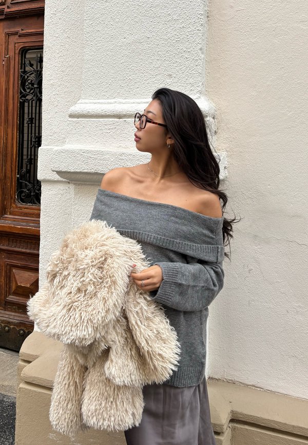 Vrouw met lang donker haar en bril, gekleed in een grijze trui met blote schouders, draagt een grote, pluizige beige jas en staat tegen een witte muur.