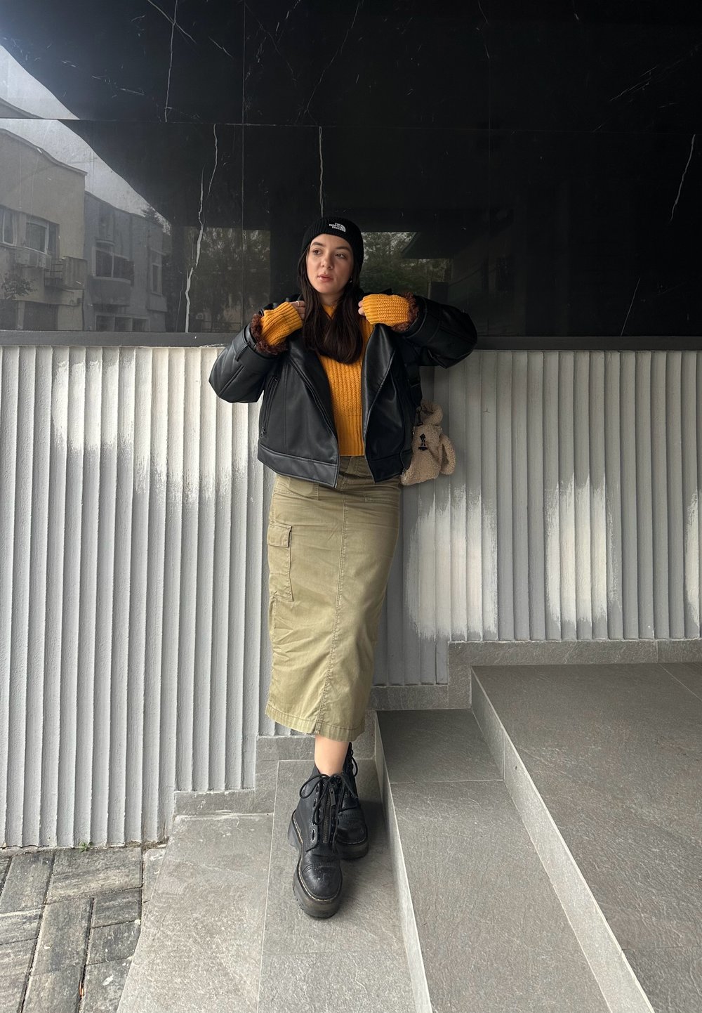 Μαύρο δερμάτινο cropped μπουφάν πάνω από κίτρινο πουλόβερ, ελαιόκαφη midi φούστα και μαύρες lace-up μπότες, στέκεται σε γκρι πέτρινες σκάλες.