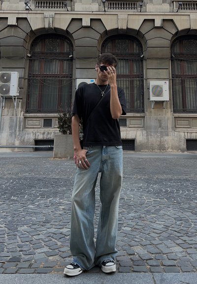Camisa negra de manga corta, jeans de pierna ancha azul claro con un lavadado degradado, gafas de sol negras, joyería de plata, de pie en una calle de adoquines.