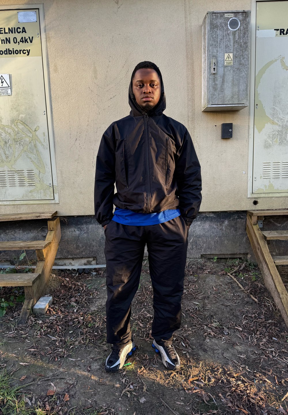 Zwarte kapjas en broek met elastische manchetten, gedragen over een blauw shirt, gecombineerd met zwarte en grijze sneakers. Op de achtergrond zijn nutsdozen te zien.