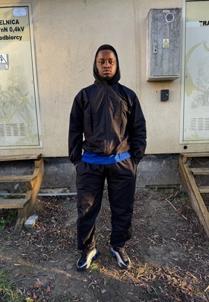 Chaqueta negra con capucha y pantalones con puños elásticos, superpuestos sobre una camisa azul, combinados con zapatillas negras y grises. El fondo muestra cajas utilitarias.