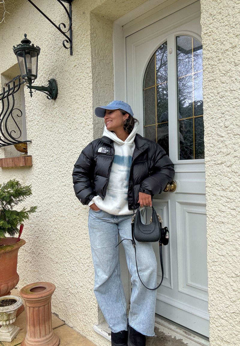 Woman in black puffer jacket and blue cap leaning against white door, holding black handbag, standing on beige porch with potted plants nearby.