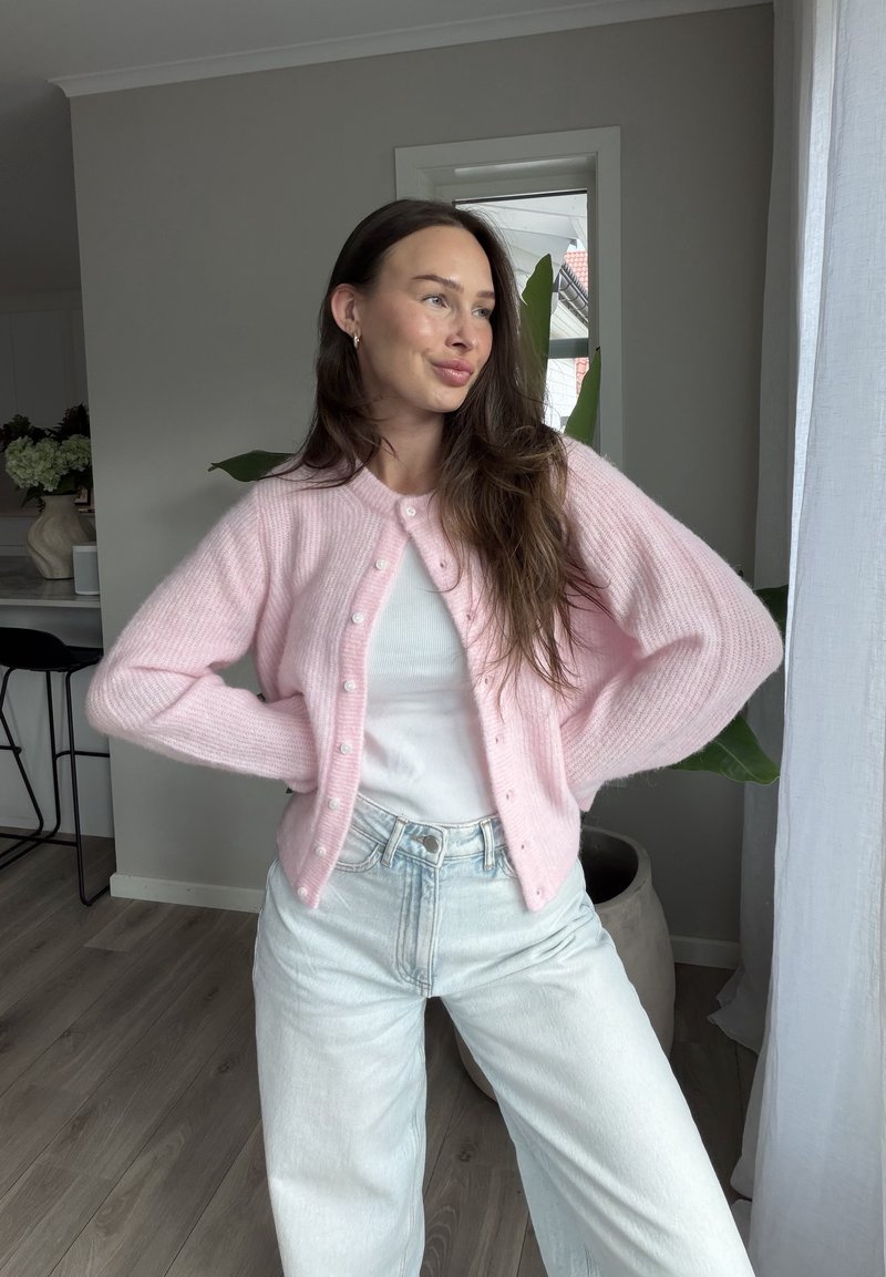 Junge Frau steht drinnen mit Händen in die Hüften gestützt, trägt eine pinke Strickjacke, ein weißes Oberteil und helle blaue Jeans, mit langen braunen Haaren.