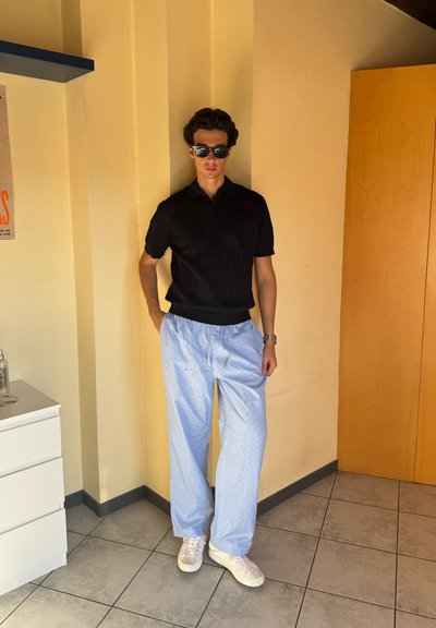 Camiseta polo negra acanalada, pantalones sueltos de rayas azul claro y zapatillas blancas, con un fondo de pared neutra y un armario de madera.
