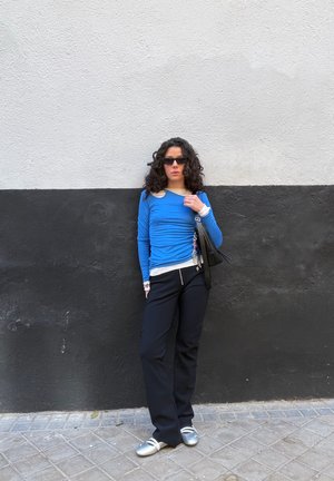 Mujer con cabello rizado que lleva gafas de sol negras, top azul de manga larga, pantalones negros y zapatos plateados apoyada contra una pared bicolor en negro y blanco.