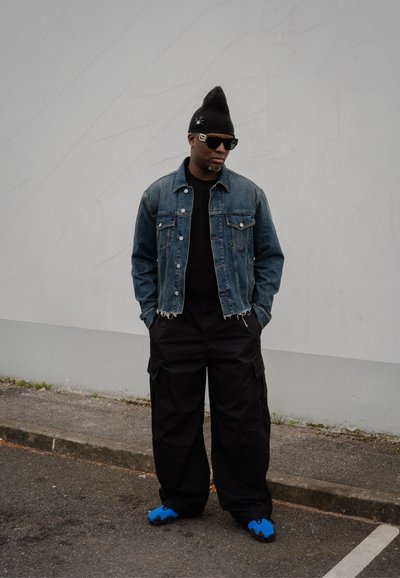 Chaqueta de denim con dobladillo desgastado, camiseta negra, pantalones cargo negros de gran tamaño y calzado azul. Accesorios incluyen un gorro y gafas de sol.