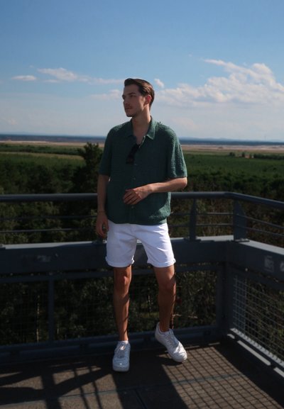 Camisa de punto verde de manga corta con cuello, pantalones cortos blancos y zapatillas blancas. Entorno al aire libre con cielo azul y campos lejanos.